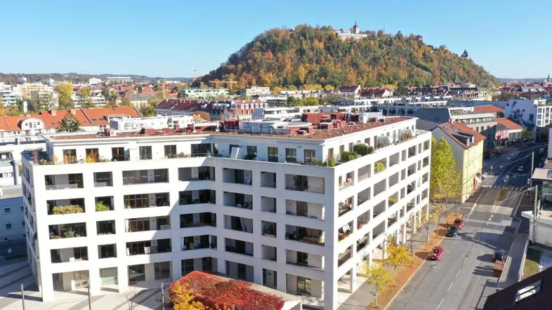 Luftaufnahme eines modernen Gebäudes mit vielen Balkonen und Blick auf einen bewaldeten Hügel.