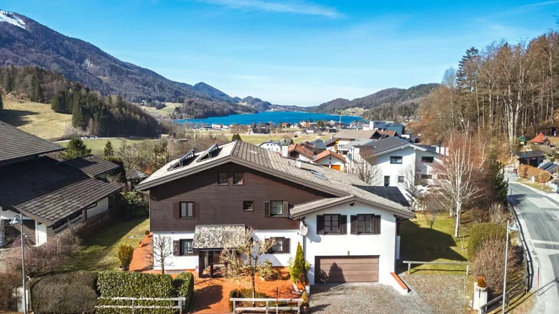 Luftaufnahme eines Hauses mit Seeblick und umliegender Berglandschaft unter blauem Himmel.