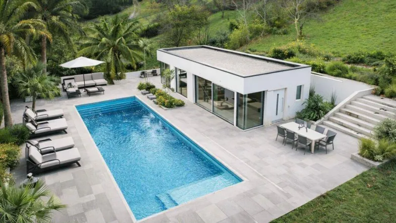 Moderne Villa mit großem Swimmingpool, Sonnenterrasse und üppiger Vegetation in einer hügeligen Landschaft.