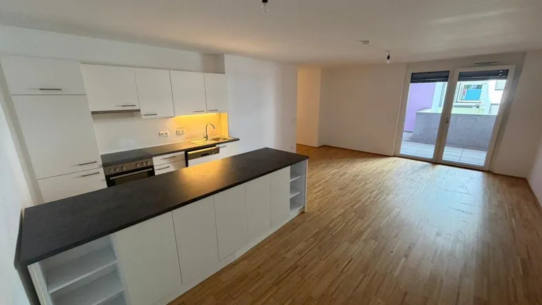 Heller offener Wohn-Essbereich mit moderner Einbauküche, Kücheninsel und Zugang zum Balkon.