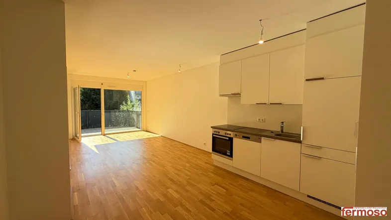 Heller offener Wohnbereich mit integrierter Küchenzeile, Parkettboden und Zugang zu einem Balkon.
