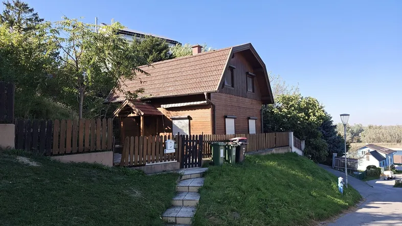 Charmantes Holzhaus auf einem Hanggrundstück mit Satteldach und umzäuntem Vorgarten unter blauem Himmel.