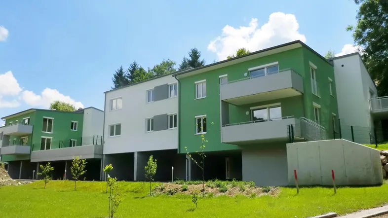 Modernes Mehrfamilienhaus mit grüner und weißer Fassade, umgeben von gepflegten Grünflächen und Bäumen.