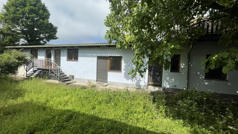 Äußere Ansicht eines älteren Hauses mit verwildertem Garten und Außentreppe unter bewölktem Himmel.