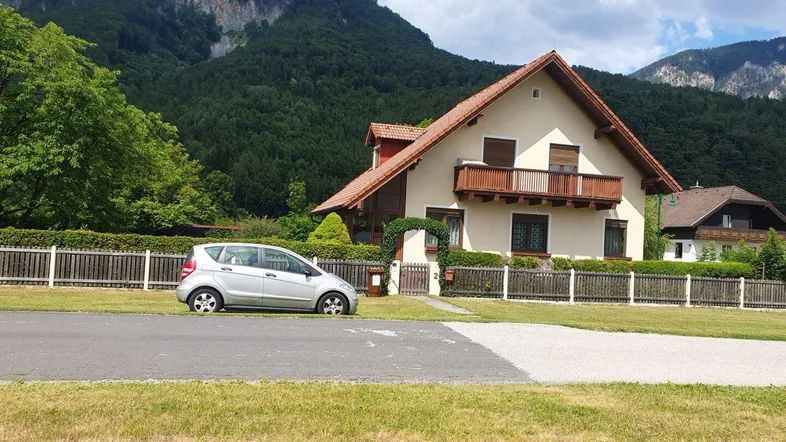 Traditionelles Haus mit Satteldach und Balkon, umgeben von grünen Bergen und einem gepflegten Vorgarten.