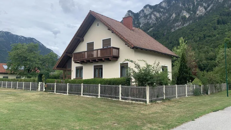 Haus mit Satteldach und Balkon vor einer beeindruckenden Bergkulisse und grünem Vorgarten.