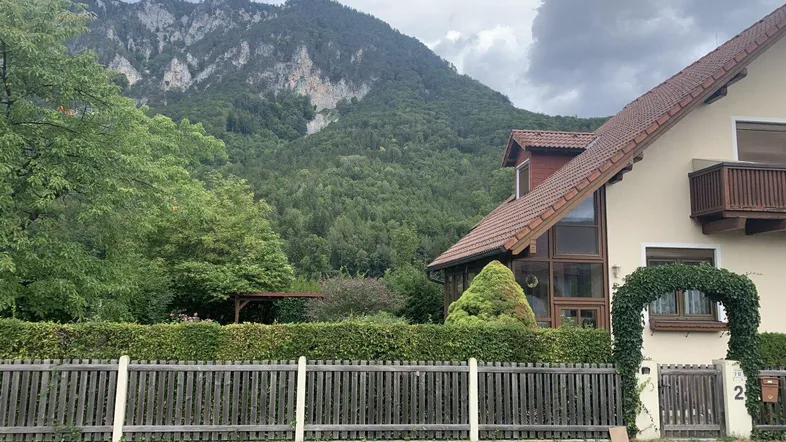 Traditionelles Haus mit Satteldach und Bergblick, umgeben von einem gepflegten Garten und Zaun.