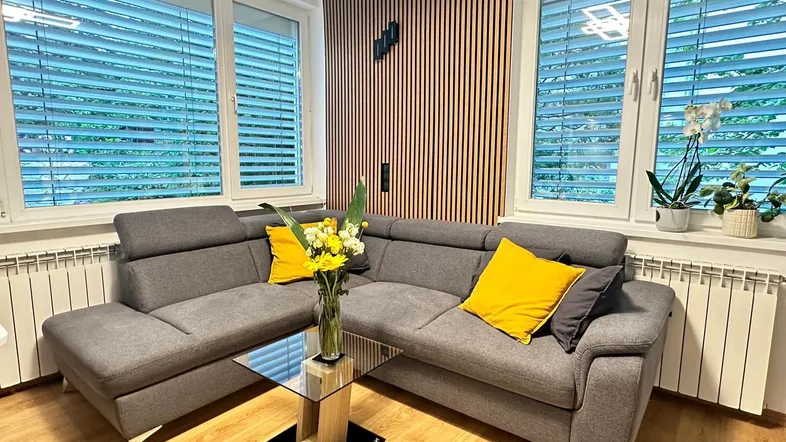 Gemütliches Wohnzimmer mit modernem Ecksofa, Holzwandverkleidung und großen Fenstern mit Jalousien.