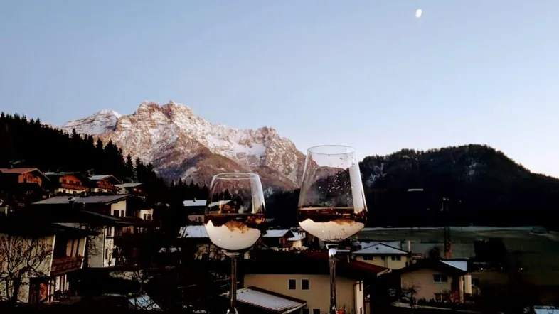 Abendstimmung mit zwei Gläsern auf einem Balkon und malerischem Blick auf schneebedeckte Berge und ein Dorf.