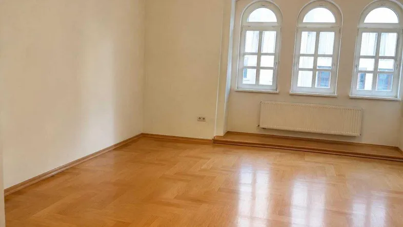 Helles Zimmer mit Fischgrätparkett, cremefarbenen Wänden und drei eleganten Rundbogenfenstern.