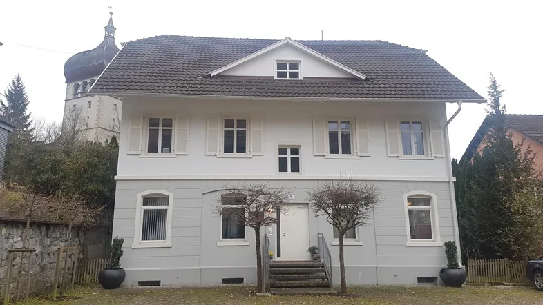 Klassisches Wohnhaus mit heller Fassade, Fensterläden und einem Kirchturm im Hintergrund.