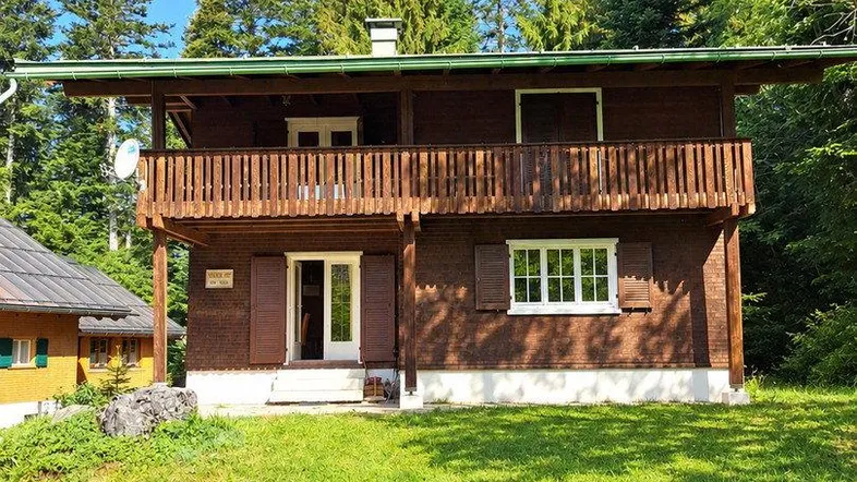 Traditionelles Holzhaus mit Balkon und grüner Umgebung, umgeben von hohen Bäumen.