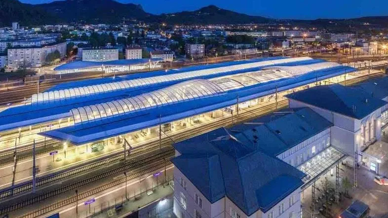 Luftaufnahme eines beleuchteten Bahnhofs und der umliegenden Stadtlandschaft bei Nacht mit Bergen im Hintergrund.
