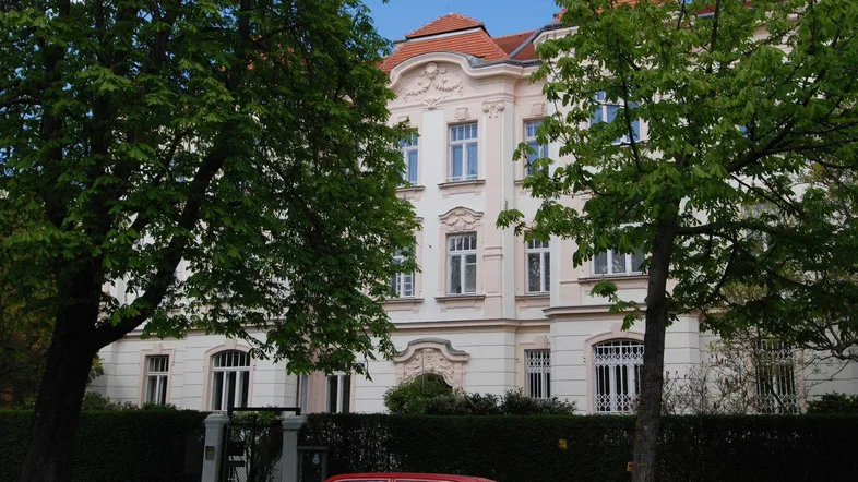 Elegantes Altbaugebäude mit verzierter Fassade, gepflegter Hecke und Bäumen im Vordergrund.