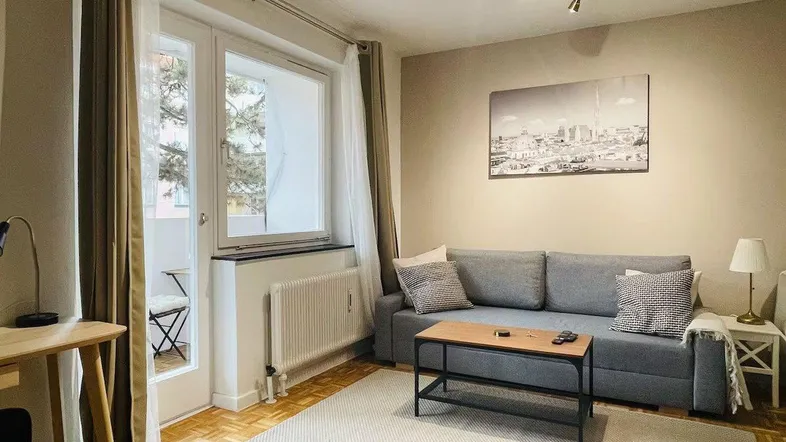 Helles Wohnzimmer mit beigem Teppich, grauem Sofa und Zugang zum Balkon.