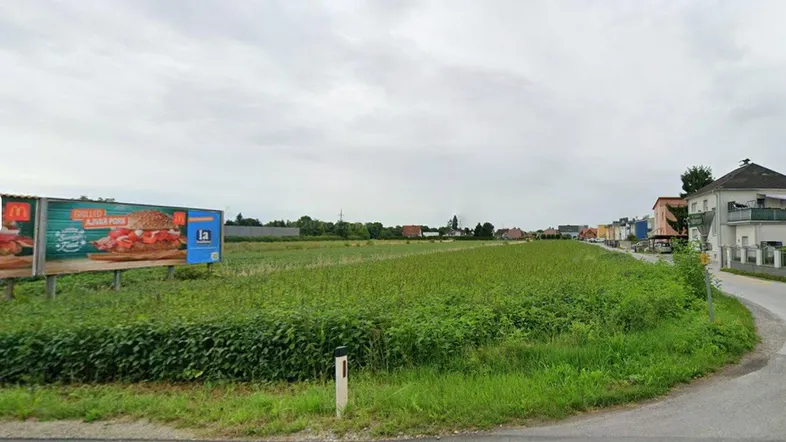 Weitläufiges grünes Feld neben einer Straße mit Gebäuden im Hintergrund und einer Werbetafel.