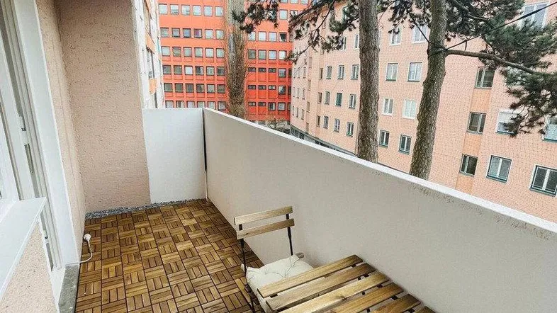 Gemütlicher Balkon mit Holzbodenfliesen und Sitzgelegenheiten, umgeben von Bäumen und Gebäuden.