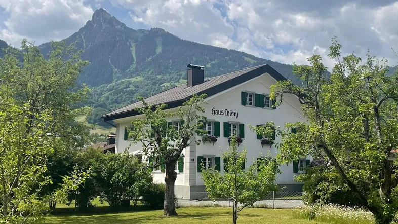 Weißes Haus mit grünen Fensterläden und Garten, umgeben von Bergen unter bewölktem Himmel.