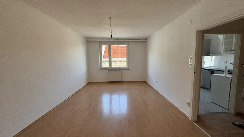 Helles, leeres Zimmer mit Parkettboden und einem Fenster, das einen Blick auf ein rotes Ziegeldach bietet.