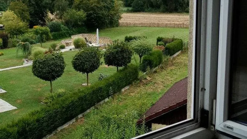 Blick aus einem geöffneten Fenster auf einen gepflegten Garten mit Hecken und weiten Feldern im Hintergrund.