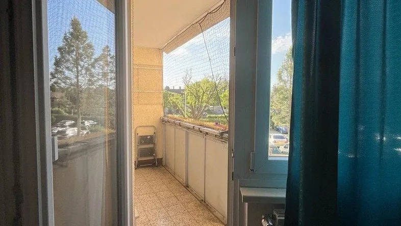 Blick aus einem Innenraum auf einen gefliesten Balkon mit Sicherheitsnetz und grüner Umgebung.
