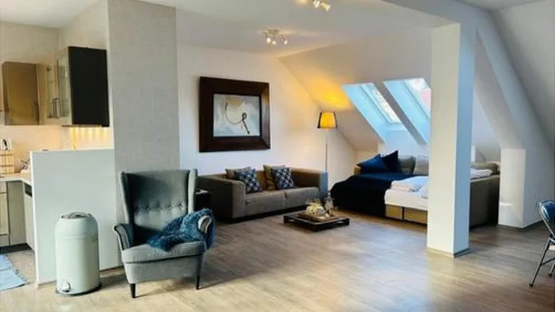 Helles Dachgeschoss-Wohnzimmer mit Holzboden, gemütlicher Sitzecke und integrierter Küchenzeile.