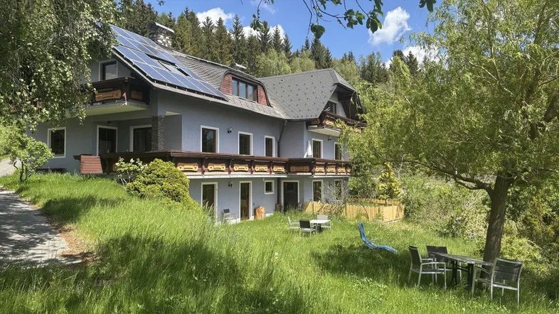 Haus mit Solaranlage, Balkonen und Gartenmöbeln in einer grünen Umgebung mit Bergblick.