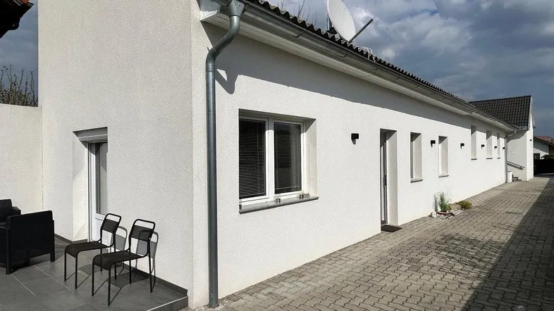 Moderne weiße Hausfassade mit kleiner gepflasterter Terrasse und Sitzgelegenheiten im Freien.