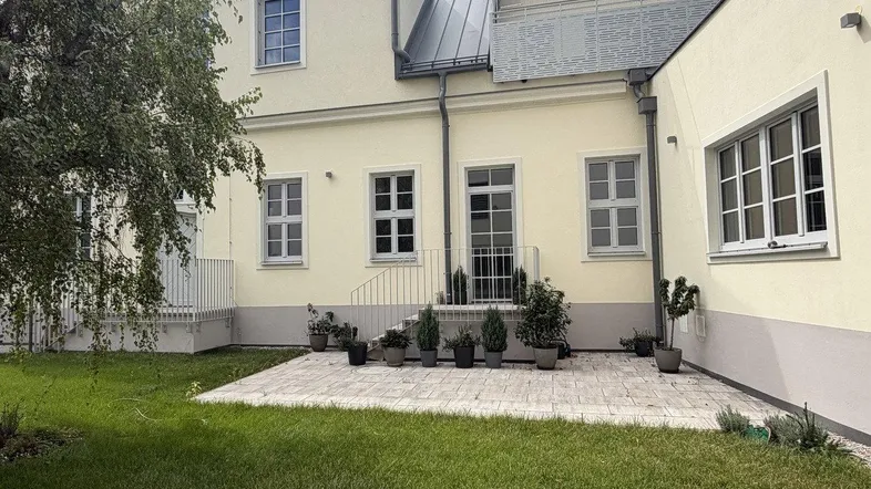 Hinteransicht des Gebäudes mit gepflegtem Rasen, gepflasterter Terrasse und kleinen Pflanzen.