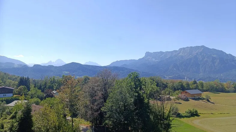 Weitläufiger Blick auf die umliegende Berglandschaft und grüne Felder unter blauem Himmel.
