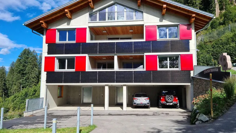 Modernes Mehrfamilienhaus mit roten Fensterläden, Solarpaneelen und Bergblick in grüner Umgebung.