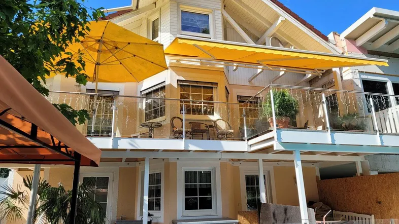 Charmantes Haus mit Balkon, gelber Markise und Sonnenschirm, umgeben von üppigem Grün.