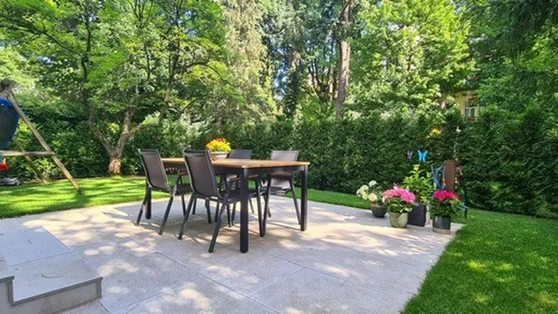 Große gepflasterte Terrasse mit Esstisch und Stühlen, umgeben von einem üppigen grünen Garten mit Schaukel.
