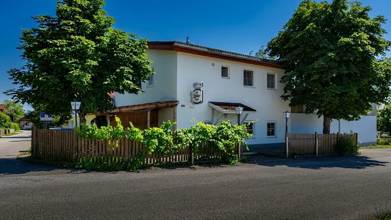 Weißes Gebäude mit grünen Bäumen und einem Holzzaun an der Straße unter blauem Himmel.