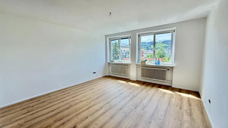 Helles Zimmer mit Laminatboden und Blick ins Grüne, ausgestattet mit Heizkörpern unter den Fenstern.