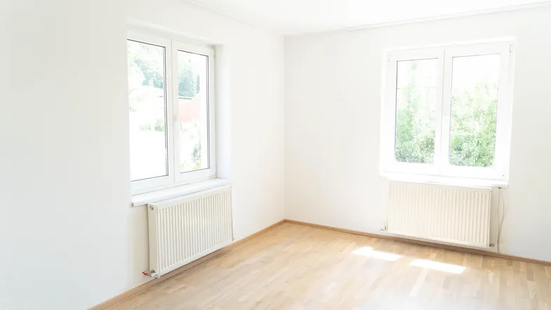 Helles, leeres Zimmer mit zwei Fenstern, Parkettboden und Heizkörpern, bereit zur individuellen Gestaltung.