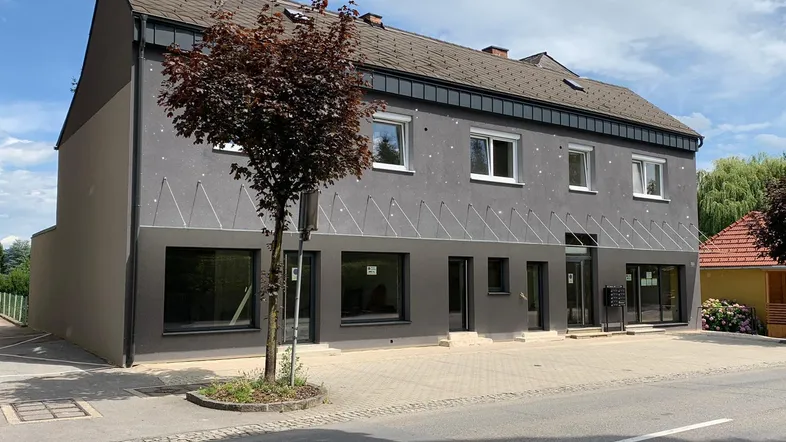 Graues Mehrfamilienhaus mit Satteldach und kleinen Bäumen vor der modernen Fassade.