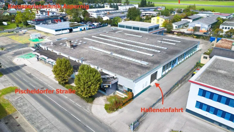 Luftaufnahme eines Gewerbegebäudes mit Flachdach, umgeben von Straßen und anderen Gebäuden, mit Bergen im Hintergrund.