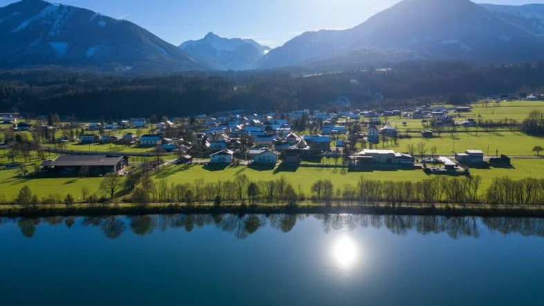 Malerische Luftaufnahme eines Dorfes am Seeufer mit majestätischen Bergen im Hintergrund unter strahlendem Sonnenschein.
