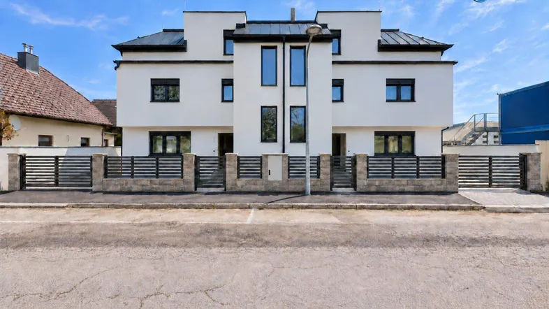 Moderne Doppelhaushälfte mit weißer Fassade, schwarzen Fenstern und Zaun an der Straße.