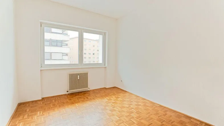 Helles Zimmer mit Parkettboden und Blick auf umliegende Gebäude, ausgestattet mit einem Heizkörper.
