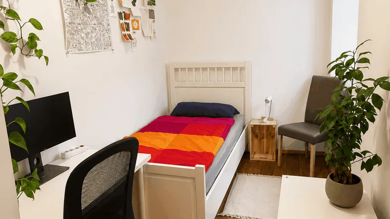 Helles Schlafzimmer mit Holzboden, Einzelbett und integriertem Arbeitsbereich am Fenster.