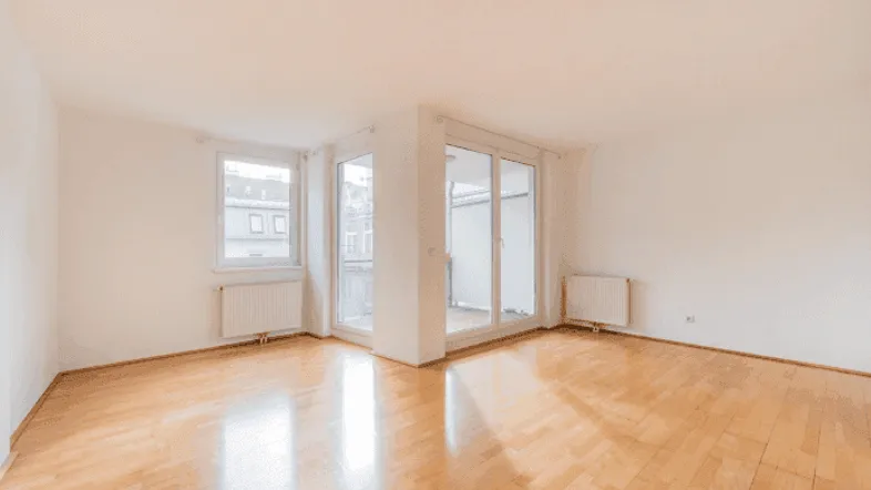 Helles, leeres Wohnzimmer mit Parkettboden und Zugang zu einem Balkon oder einer Terrasse.