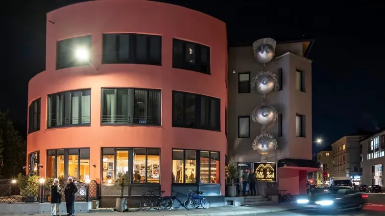 Modernes Gebäude mit runder Fassade und auffälliger Kunstinstallation bei Nachtbeleuchtung.