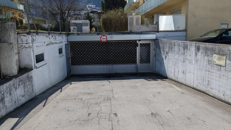 Einfahrt zu einer Tiefgarage mit Metalltor und angrenzendem Wohngebäude in bergiger Umgebung.