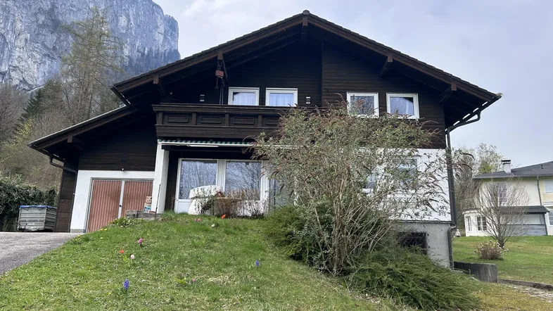 Dunkles Holzhaus mit Satteldach und Balkon, eingebettet in eine grüne Landschaft mit Bergblick.