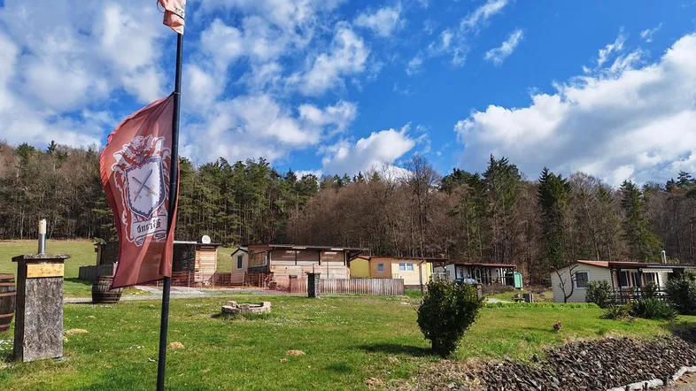 Ländliche Ansicht mit mehreren kleinen Hütten, grüner Wiese und Wald im Hintergrund unter blauem Himmel.
