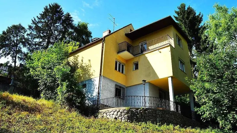 Gelbes Haus mit Balkonen und einer Terrasse, umgeben von viel Grün und Bäumen.