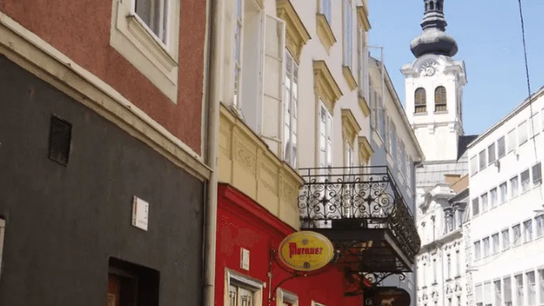 Historische Gasse mit traditionellen Gebäuden, einem Balkon und einem Kirchturm im Hintergrund.