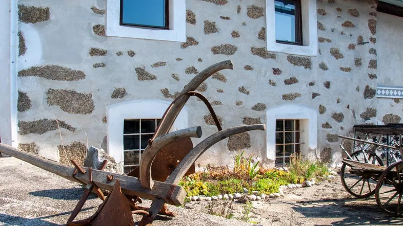 Altes Bauernhaus mit rustikaler Steinfassade und historischen landwirtschaftlichen Geräten im Vorgarten.